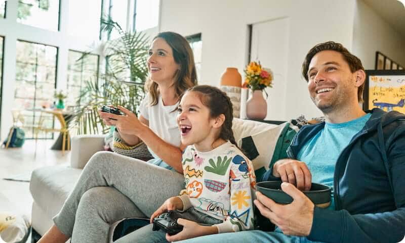 A mother and daughter are sitting on a couch, playing video games together while the father watches and eats popcorn