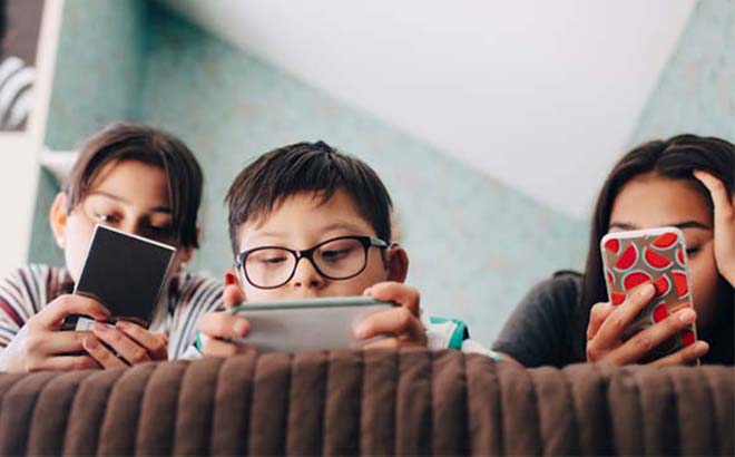 Three kids lounging on a sofa and browsing the web on their respective devices.